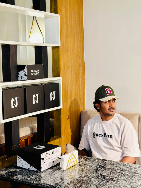 A man wearing a white t-shirt with 'Question' printed on the chest and a dark green cap sits in a modern room next to a marble table, with branded shoe boxes on display.