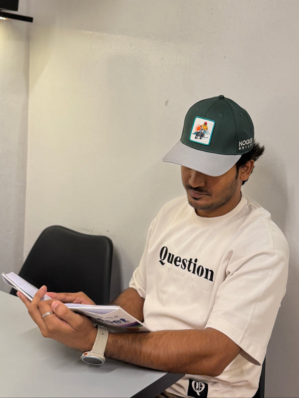 A man wearing a green and white baseball cap and a cream-colored t-shirt with the word 'Question' printed on the chest, sitting at a table and reading a document.