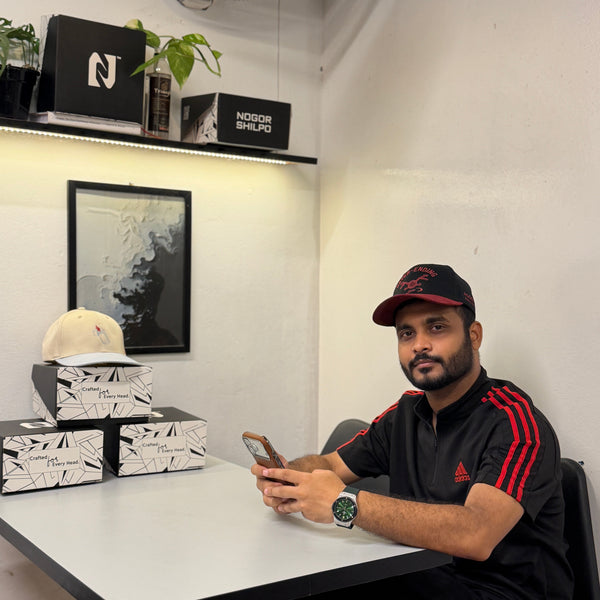 A man wearing a black Adidas track jacket with red stripes and a matching cap sits at a white table, holding a phone. Several branded black and white boxes are stacked on the table next to him, along with a small plant and framed wall art in a home office setting.